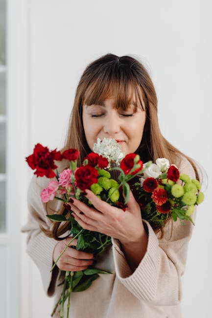 A woman with long brown hair and closed eyes gently holding a vibrant bouquet of fresh flowers, including red carnations, pink roses, white carnations, and green chrysanthemums, wrapped in light brown