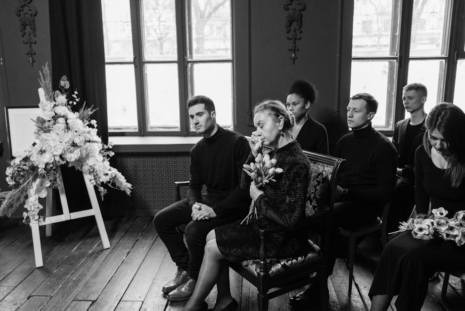 A woman holding a bouquet of fresh flowers, including tulips and roses, seated in a black patterned dress in a solemn setting. She is surrounded by five young adults, all dressed in dark clothing, wit