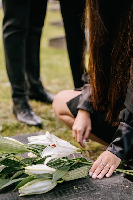 A person in black attire with long brown hair is kneeling beside a gravestone, carefully placing a white lily flower with its petals slightly open and green leaves surrounding it, as part of a floral