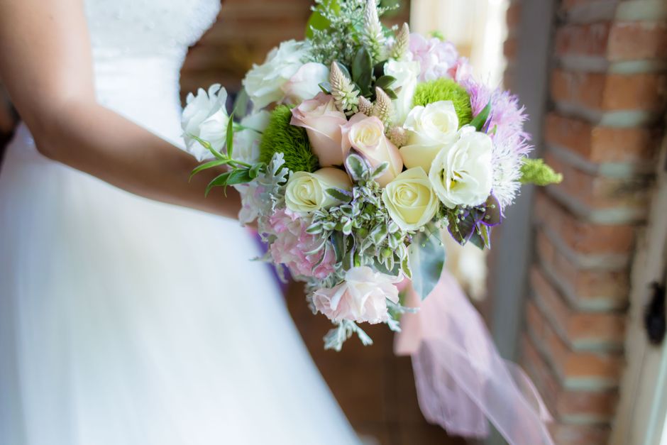 A bride in a white wedding dress holds a lush bouquet consisting of white roses, light pink roses, and pale green flowers, accented with lavender and silver-green foliage. The bouquet is wrapped with 