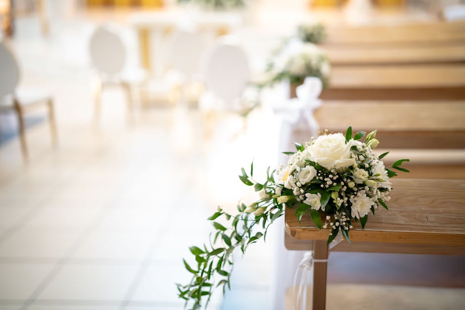A close-up of a wedding floral arrangement placed on a wooden pew in a softly lit indoor ceremony space. The bouquet features a large white rose surrounded by smaller white flowers such as spray roses