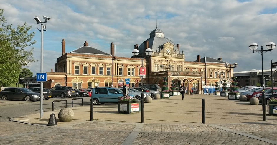 A photograph of Isleworth High Street showing a historic red brick building with a central clock tower and arched windows, set against a partly cloudy sky. In the foreground, there is a spacious paved