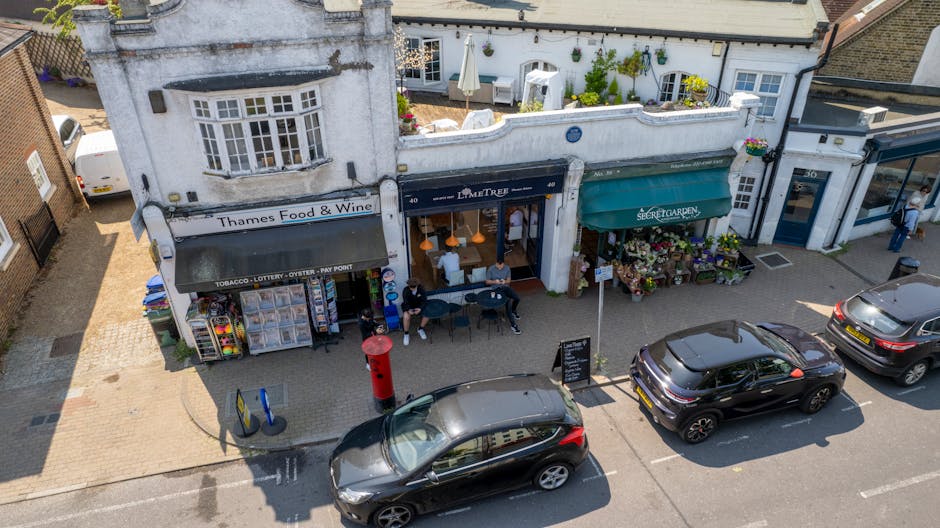 An aerial view of a row of small shops on Isleworth High Street, featuring a floral shop with a green awning named 'Secret Garden' displaying potted plants and flowers outside. Next to it, a café has