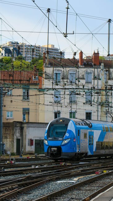 A modern blue and silver train traveling on railway tracks in an urban setting, surrounded by overhead electric wires and a background of multi-story buildings with tiled rooftops and chimneys. The sc