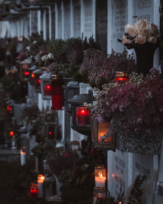 A memorial wall decorated with numerous floral arrangements and candles, featuring a variety of flowers including pale pink roses, white carnations, and purple statice, arranged in hanging and standin