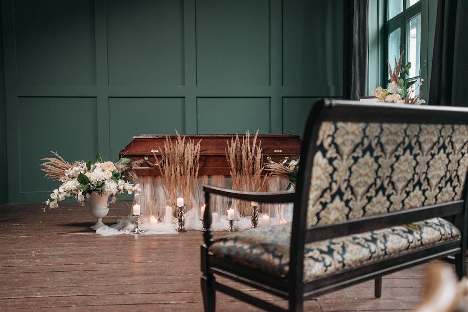 A floral arrangement featuring white and blush pink flowers, including roses and peonies, complemented by greenery, arranged in a white ceramic vase. The flowers are positioned on a wooden floor again